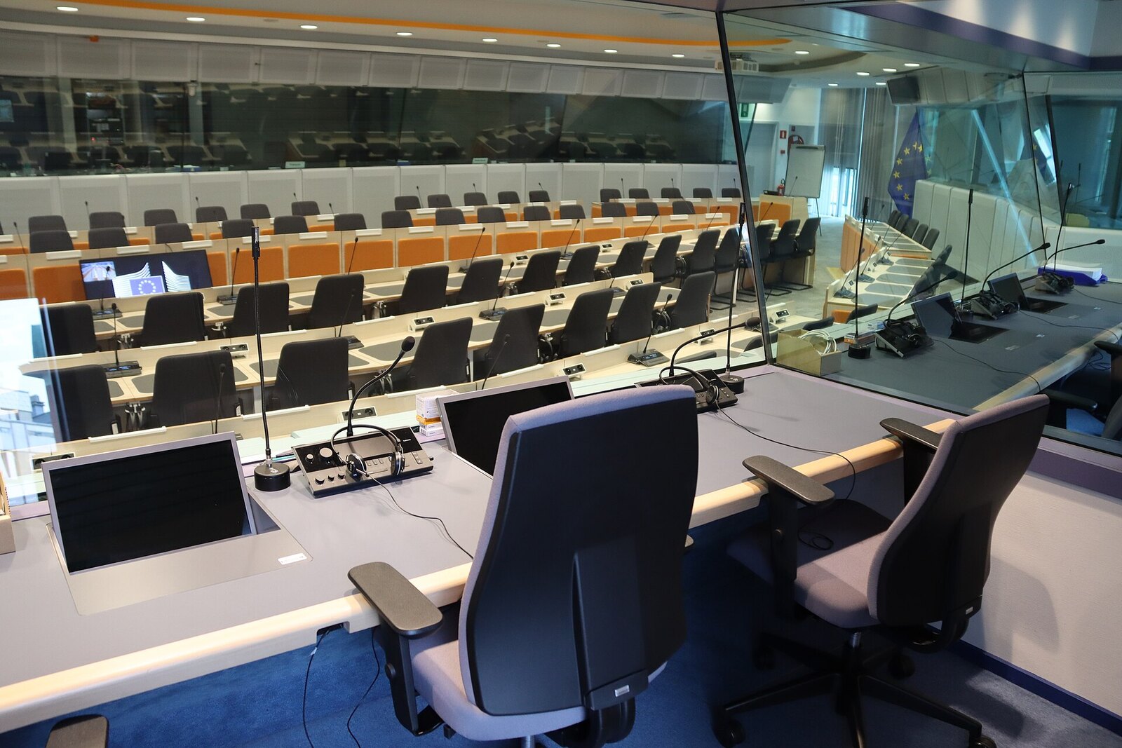 An empty European Union committee room in Brussels seen from an interpreter's booth, rows of desks with microphones and an EU flag
